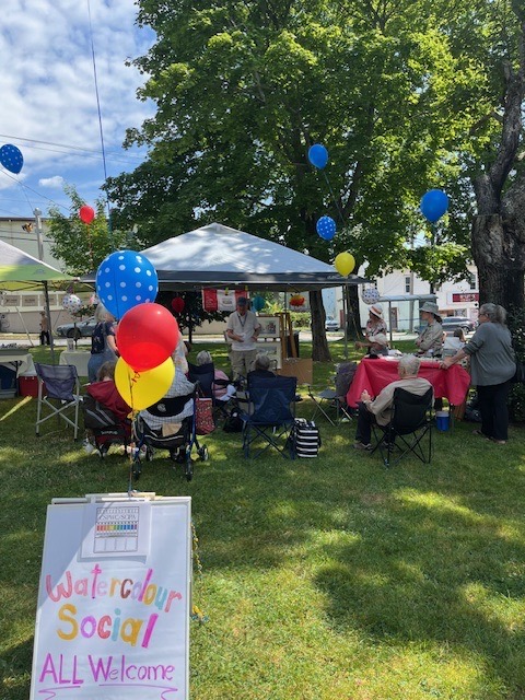 Shows a watercolour social gathering at Hydrostone Park in Halifax, NS. There is a an awning covering a table, people seated in lawn chairs and balloons attached to a sign that says “watercolour social”
