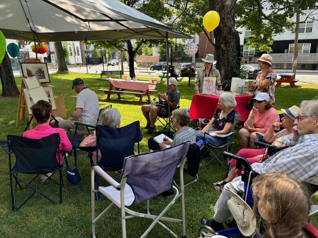 Shows an artist at his easel with onlookers seated where they can watch him paint. This is outdoors at Hydrostone Park in Halifax, NS