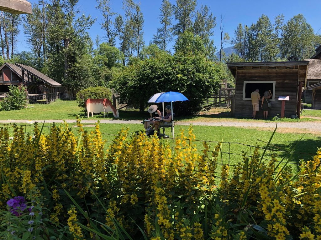 Artists at work at their easels, seated under umbrellas, on a green lawn on a sunny day. At Kilby Historic Village